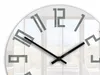 A close-up of a modern wall clock with a minimalist design, showing the numbers and hands against a blurred background of a room with windows. Produktbild-Vorschau