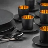A set of black espresso glasses with gold interiors and black saucers are arranged on a dark gray surface, with a fork and spoon in the foreground. Náhled obrázku produktu