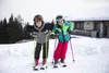 Two children in ski gear posing in the snow with skis and poles, a building in the background. Produktbild-Vorschau