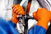 A close-up shot of two hands wearing orange nitrile gloves, turning a metal valve on a large industrial tank. Náhľad obrázku produktu