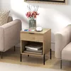 A bedside table with a drawer and open shelf is placed next to a sofa in a living room setting. A vase of flowers, a phone, and a cup of coffee are on top of the table. Produktbild-Vorschau