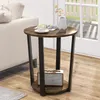 A round side table with a rustic brown finish and black metal legs is placed next to a sofa. The table has a lower shelf with a small radio on it. A vase with branches and a cup of coffee are on the tabletop. Náhled obrázku produktu