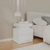 A white nightstand with two drawers is placed next to a bed in a bedroom. The nightstand has a lamp, a clock, and some books on top. Produktbild-Vorschau