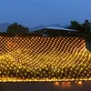 A net of yellow lights is draped over bushes at dusk. Produktbild-Vorschau