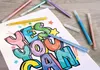 A coloring page with the words "YES YOU CAN" written in large, 3D-style letters, being colored in with glitter pens. Several glitter pens are scattered around the page and on a wooden surface. Podgląd zdjęcia produktu
