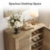 A close-up view of a wooden shoe cabinet with its doors open, displaying shoes on shelves. The top surface is decorated with a vase of flowers, candles, and books, with a lamp in the background. Produktbild-Vorschau
