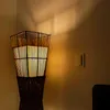 A floor lamp with a woven bamboo and rattan shade is illuminated, casting shadows on a light-colored wall. A light switch is visible on the wall to the right. Produktbild-Vorschau