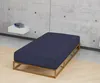 A navy blue fitted sheet on a wooden bed frame against a concrete wall. Produktbild-Vorschau