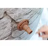 A brown skimmer with an inlet nozzle is attached to a wall with a stone pattern. A hand is blurred in the background, reaching towards the product. Produktbild-Vorschau
