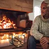 A man sits in a chair next to a fireplace, with a log holder in the foreground. Náhled obrázku produktu