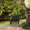 A black hanging chair with a pillow is suspended from a tree branch in a garden setting. Voir les images du produit