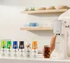 A person is holding a glass of soda next to a SodaStream machine and several flavored syrup bottles. Podgląd zdjęcia produktu