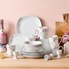 A collection of white ceramic dinnerware with a pink floral pattern, arranged on a wooden surface against a light pink background. Voir les images du produit