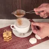 A person is dusting cinnamon onto a cappuccino with a sieve, with cookies and meringues on the side. Náhled obrázku produktu