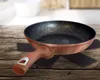 A copper-colored frying pan with a wooden handle is placed on a wooden table, with black placemats underneath. Voir les images du produit