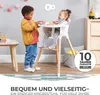 Two young children are playing with blocks near a high chair. One child is standing and handing blocks to the other child who is sitting in the high chair. Produktbild-Vorschau