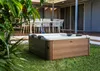 A hot tub is placed on a grassy lawn in front of a patio with white chairs and a table. Náhled obrázku produktu