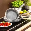 A close-up shot of two stainless steel frying pans on a stovetop, with cherry tomatoes on a plate in the foreground and potted herbs and lemons in the background. Náhľad obrázku produktu