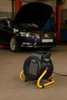 A black and yellow electric heater is placed on the floor in front of a car with its hood open in a garage. Voir les images du produit