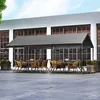 An outdoor seating area with tables and chairs under a gray canopy, next to a modern building with large windows. Produktbild-Vorschau