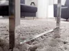 A close-up shot of the chrome leg of a coffee table resting on a plush, grey rug. In the background, a dark grey sofa and armchair are visible, suggesting a living room setting. Produktbild-Vorschau