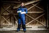 A man in workwear stands in front of a rustic wooden door. Voir les images du produit