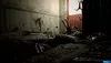 A dark, unsettling scene with what appears to be a body or bodies covered in a dark, viscous substance, possibly in a dilapidated room with a red door in the background. Produktbild-Vorschau