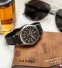 A watch, wallet, and perfume bottle are arranged on a white surface, with sunglasses in the background. Produktbild-Vorschau