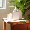 A white tissue box shaped like a house sits on a wooden table next to a glass of iced drink, with a large green plant in the background. Anteprima dell’immagine prodotto