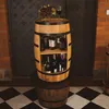 A wooden barrel-shaped mini bar shelf filled with bottles of alcohol and topped with grapes and a glass of wine, placed on a checkered floor in front of a brick wall and a dark metal door. Náhled obrázku produktu