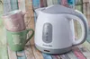 A white and gray electric kettle sits next to two stacked mugs on a colorful wooden background. Produktbild-Vorschau