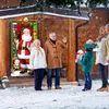 A family is receiving Christmas gifts from Santa Claus in front of a decorated house during a snowy day. Produktbild-Vorschau