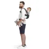 A man is carrying a baby in a carrier on his back, holding a teddy bear in his arm. The background is white. Náhled obrázku produktu