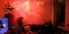 A person is sitting on a couch in a room illuminated by a star projector, creating a galaxy effect on the walls and ceiling. Podgląd zdjęcia produktu