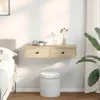 A floating shelf with two drawers is mounted on a white wall above a round white stool. A bed is partially visible on the left, and a large plant is on the right. Náhled obrázku produktu