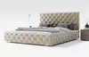 A tufted beige bed with pillows and a bedside table with a lamp and vase. Náhled obrázku produktu