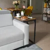 A C-shaped side table is placed next to a sofa, holding a cup of coffee, a croissant, and a vase with flowers. In the background, a dining table with chairs and a shelf unit are visible. Produktbild-Vorschau