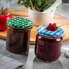 Two jars of preserves or jam with checkered lids, one topped with red currants, sit on a countertop next to a tomato and a mortar and pestle with herbs. Produktbild-Vorschau
