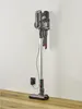 A cordless vacuum cleaner is shown standing upright, charging on a wall-mounted dock connected to an electrical outlet. The vacuum is a 2-in-1 handheld and stick model with a gray and silver body and red accents. It is positioned on a wooden floor against  Produktbild-Vorschau