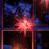 A red star-shaped Christmas light hangs in a window at night, with a blurry background of a room with a fireplace. Produktbild-Vorschau