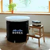 A portable bathtub filled with water, placed on a rug next to a wooden stool with folded towels and bottles. Produktbild-Vorschau