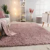 A fluffy pink rug is laid out in a living room setting, with a sofa, a floor lamp, and a shelf unit visible. Produktbild-Vorschau
