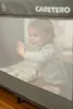 A baby is sitting inside a travel crib, looking out and waving. Anteprima dell’immagine prodotto