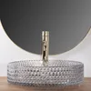 A glass vessel sink with a textured pattern and a gold faucet in front of a large round mirror. Anteprima dell’immagine prodotto