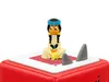 A small figurine of a cartoon character sits on top of a red cube-like object, with two gray shark fins protruding from the surface it sits on. Produktbild-Vorschau