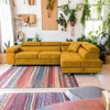 A yellow sectional sofa is placed in a room with many plants and colorful rugs. Produktbild-Vorschau