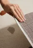 A hand is shown lifting the edge of a rug, revealing its backing. Anteprima dell’immagine prodotto