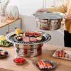 A person is holding a grill lid over a BBQ grill with food on it, set on a wooden table with various food items around. Náhled obrázku produktu