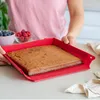 A person is holding a red baking pan with a cake inside, with bowls of berries in the background. Produktbild-Vorschau