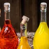 Three glass bottles filled with colored liquids, two red and one yellow, are arranged closely together. The middle bottle's cork is tilted and partially out of the neck. The background is a dark, rustic wooden surface. Podgląd zdjęcia produktu